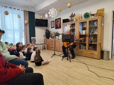 Mężczyzna siedzi przy drewnianej szafie i gra na gitarze przy mikrofonie. Grupa kilkunastu dzieci siedzi na podłodze po lewej i uważnie słucha. Wewnątrz pomieszczenia wiszą złote balony i białe ozdoby papierowe. Duże okno z jasnymi zasłonami znajduje się po lewej stronie.