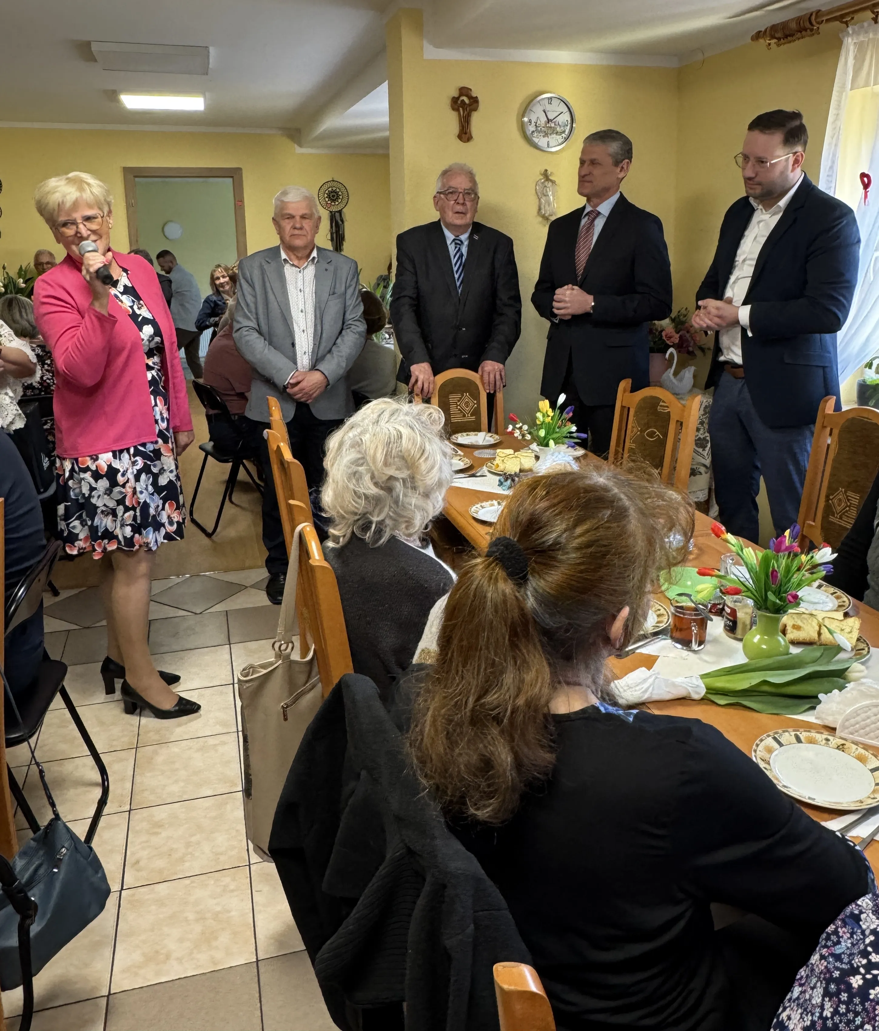 Kobieca osoba w różowej marynarce stoi po lewej i mówi do mikrofonu. Czterech mężczyzn stoi przy ścianie z zegarem i krzyżem. Przy środku stołu siedzą osoby plecami, na stole talerze, przekąski i wazoniki z kwiatami. Pomieszczenie jest wypełnione ludźmi, wnętrze ma żółte ściany i światło dzienne.