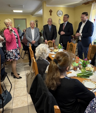 Kobieca osoba w różowej marynarce stoi po lewej i mówi do mikrofonu. Czterech mężczyzn stoi przy ścianie z zegarem i krzyżem. Przy środku stołu siedzą osoby plecami, na stole talerze, przekąski i wazoniki z kwiatami. Pomieszczenie jest wypełnione ludźmi, wnętrze ma żółte ściany i światło dzienne.