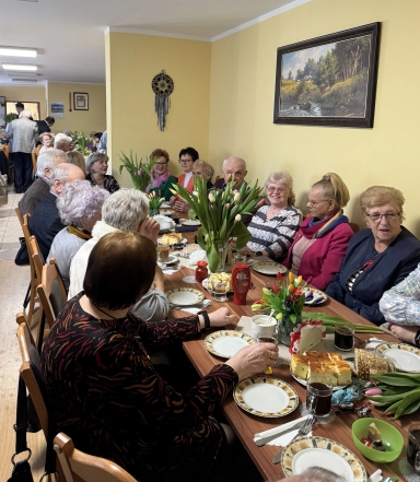 Grupa starszych osób siedzi przy długim stole w jasnym, żółtym pomieszczeniu. Na stole leżą talerze, kawa, ciasta i bukiety tulipanów w wazonach. Na ścianie wisi obraz krajobrazu i dekoracja ścienna.