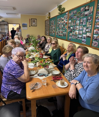 Kilkanaście starszych kobiet siedzi przy długim drewnianym stole w jasnym pomieszczeniu. Stół jest zastawiony talerzami, filiżankami, bukietami tulipanów i przekąskami. Na ścianie wisi duża tablica z licznymi drobnymi fotografiami.