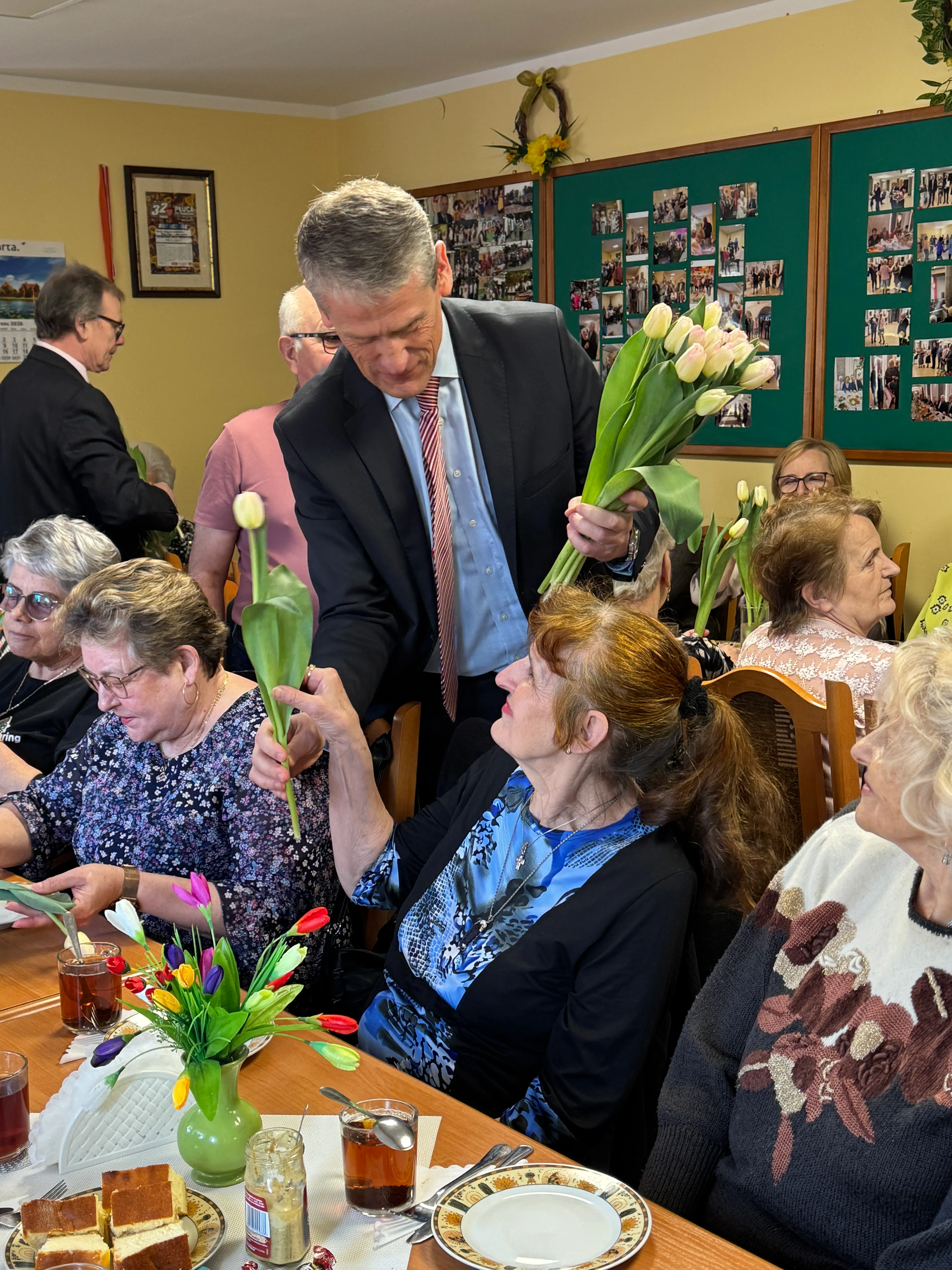 Mężczyzna w garniturze wręcza bukiet białych tulipanów starszej kobiecie przy stole. Kilka starszych kobiet siedzi wokół stołu z filiżankami herbaty i talerzami ciasta. Na stole stoi zielony wazon z kolorowymi tulipanami i słoik musztardy. Wnętrze sali z żółtymi ścianami i tablicami ze zdjęciami na ścianie.