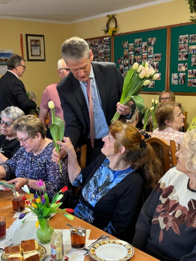 Mężczyzna w garniturze wręcza bukiet białych tulipanów starszej kobiecie przy stole. Kilka starszych kobiet siedzi wokół stołu z filiżankami herbaty i talerzami ciasta. Na stole stoi zielony wazon z kolorowymi tulipanami i słoik musztardy. Wnętrze sali z żółtymi ścianami i tablicami ze zdjęciami na ścianie.