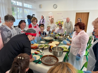 Grupa starszych osób stoi wokół stołu w jasnym pomieszczeniu, uczestnicząc w warsztatach kulinarnych. Kobieta w czarnym ubraniu i czerwonej chuście przygotowuje jedzenie na środku stołu, na którym znajdują się różne składniki i miski. Wszyscy mają założone plastikowe fartuchy i niebieskie rękawiczki. W tle widać duże okno i zegar na ścianie.