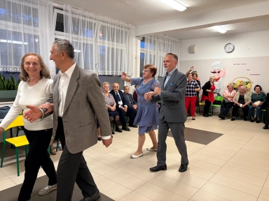 W sali z jasnymi ścianami i dużymi oknami tańczą dwie pary starszych osób, jedna para idzie na przód po lewej, druga – po prawej. W tle kilkanaście osób siedzi lub stoi przy ścianie. Na ścianie znajduje się zegar i kolorowa grafika z postacią kucharza oraz koszem owoców.