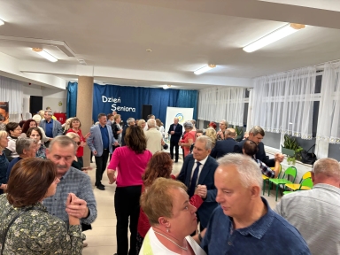 Grupa starszych osób tańczy w jasnym, dużym pomieszczeniu z białymi ścianami i oknami udekorowanymi firankami. W tle widoczna granatowa kurtyna z napisem „Dzień Seniora”. Niektóre osoby są w parach, na środku sali. Po prawej stronie widać rząd zielono-żółtych krzeseł i rośliny na parapecie.