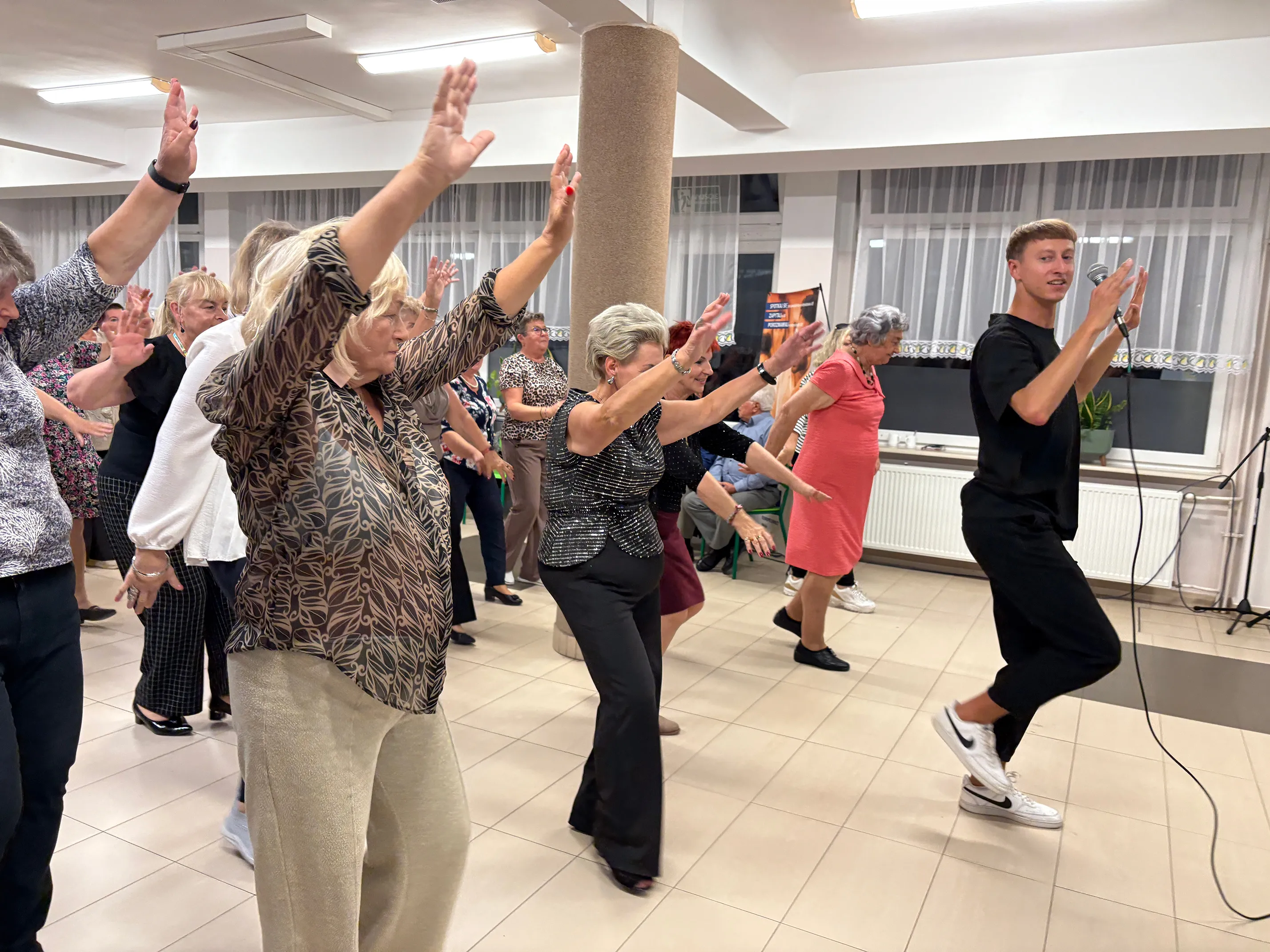 Grupa seniorów tańczy w świetle sztucznym na dużej sali, młody instruktor prowadzi zajęcia, trzymając mikrofon, wszyscy mają uniesione ręce.