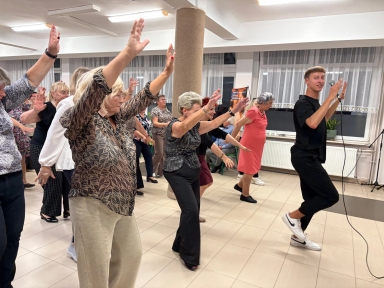 Grupa seniorów tańczy w świetle sztucznym na dużej sali, młody instruktor prowadzi zajęcia, trzymając mikrofon, wszyscy mają uniesione ręce.