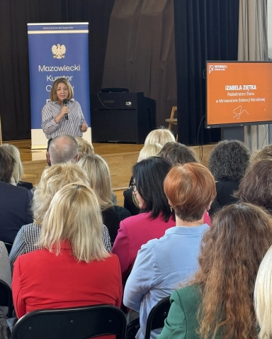 Kobieta stoi na scenie i mówi do publiczności, trzymając mikrofon. Wnętrze sali z drewnianą sceną i ciemnymi kurtynami. Po lewej stoi niebieski pionowy baner, po prawej stoi pomarańczowy ekran. Kilkadziesiąt osób siedzi przodem do sceny, widoczne tylne części głów i kolorowe ubrania.