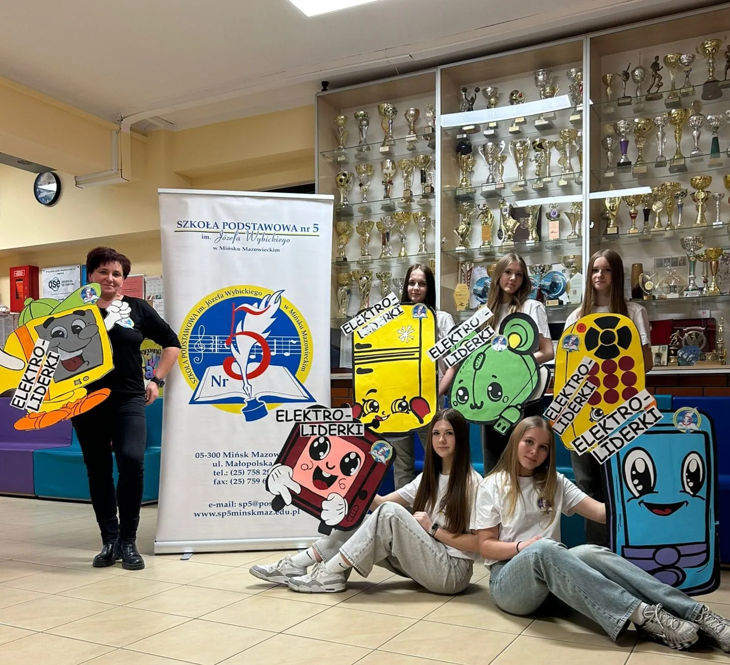 Sześć osób — jedna dorosła kobieta po lewej i pięć nastolatek — stoi i siedzi w szkolnym holu. Gablota z wieloma pucharami zajmuje tło. Pośrodku stoi stojak z napisem Szkoła Podstawowa nr 5. Uczestniczki trzymają duże, kolorowe kartonowe postacie z napisami ELEKTRO-LIDERKI.