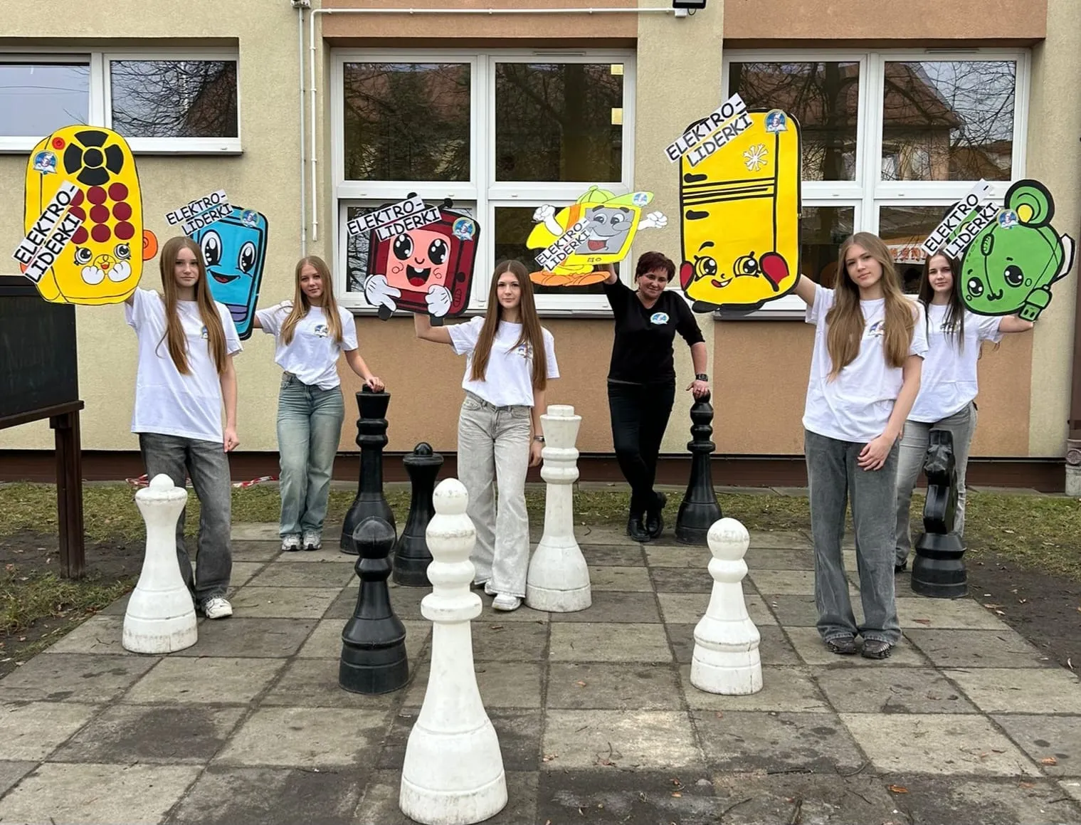 Siedem osób stoi na zewnątrz przed beżowym budynkiem z oknami. Większość nosi białe koszulki, jedna osoba ma czarną. Każda trzyma dużą, kolorową wyciętkę z rysunkową postacią przypominającą urządzenia elektroniczne z oczami. Przed nimi na bruku ustawione są duże czarne i białe figury szachowe.