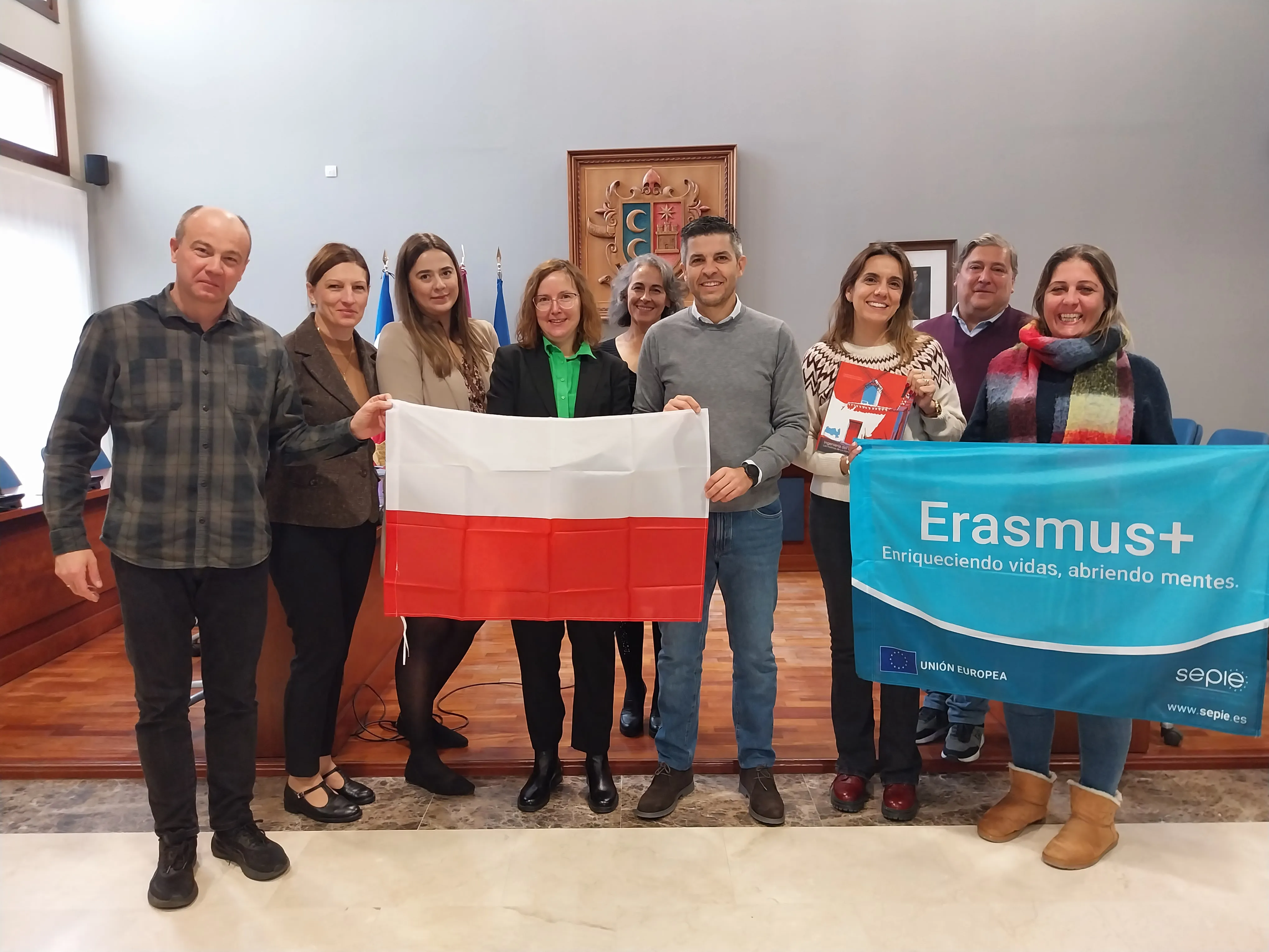 Dziewięć osób stoi w pomieszczeniu z szarą ścianą i drewnianą podłogą. Trzy osoby z przodu trzymają flagę Polski, a dwie po prawej stronie trzymają niebieski baner z napisem Erasmus+. Za nimi wisi drewniana dekoracja i kilka flag. Wszystkie osoby uśmiechają się.