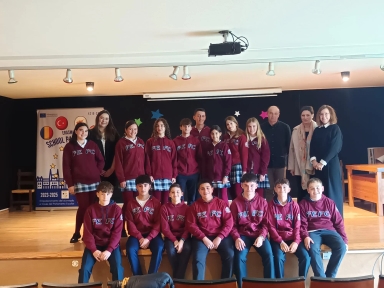 Grupa szesnastu uczniów i trzech dorosłych stoi i siedzi na scenie w szkolnej auli. Uczniowie noszą bordowe bluzy z napisem „FEFC” oraz granatowe spodnie lub spódniczki w kratę. W tle jest czarna ściana ozdobiona kolorowymi gwiazdkami oraz baner z logo Erasmus+ i flagami Rumunii i Turcji. Sala jest dobrze oświetlona światłami sufitowymi.