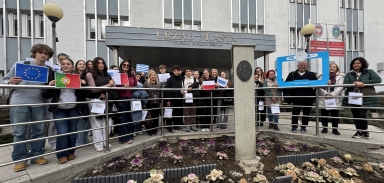 Około siedemnastu nastolatków i trzy dorosłe osoby siedzą i rozmawiają wokół dużego drewnianego stołu w jasnej sali konferencyjnej. Po prawej stoi kobieta przy ściennym monitorze z kolorowym slajdem, za grupą stoi flipchart i okno z pionowymi żaluzjami.