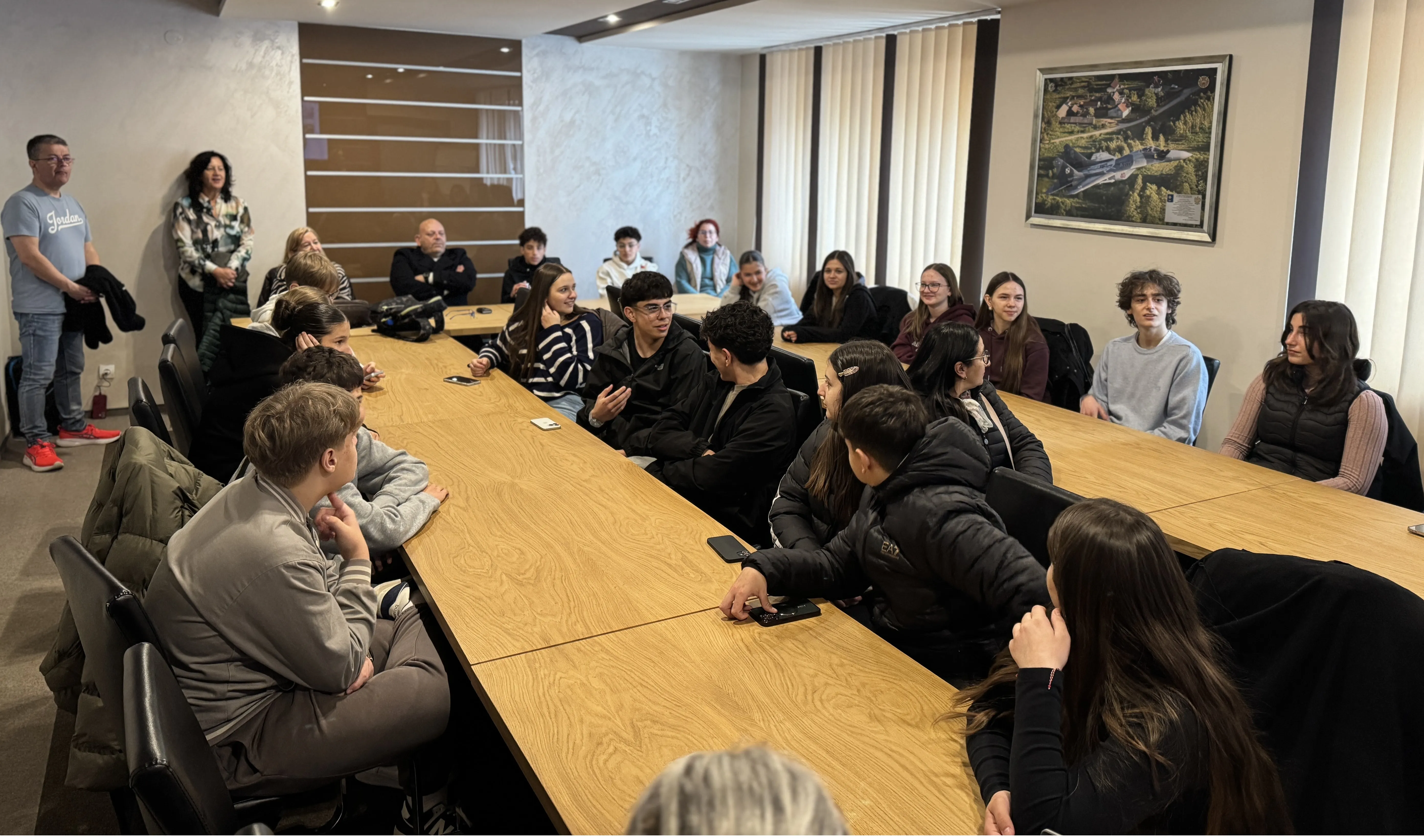 Kilkunastu młodych ludzi siedzi wokół długiego drewnianego stołu w jasnej sali konferencyjnej. Dwie osoby stoją przy ścianie z lewej strony, za stołem są pionowe żaluzje i wiszący obraz na ścianie.