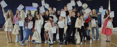 Grupa dzieci i nastolatków stoi na hali sportowej, trzymając w górze dyplomy i torby z nadrukiem roweru. W tle widoczna jest tablica informacyjna i ściana z ciemnym tłem.