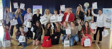 Grupa 17 osób w hali sportowej, w większości kobiet i kilka dzieci, trzyma w górze dyplomy i białe torby prezentowe, część osób klęczy, część stoi, wszyscy uśmiechają się. W tle widać plakaty i dekoracje.