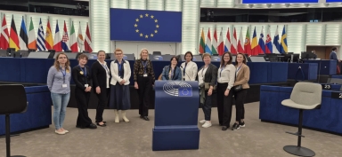 Dziesięć kobiet stoi w sali obrad z niebieskim mównicą z symbolem Unii Europejskiej na pierwszym planie. W tle znajduje się wiele flag państw członkowskich UE oraz duży niebieski baner z żółtym okręgiem gwiazd. Pomieszczenie jest nowoczesne, wewnątrz budynku.