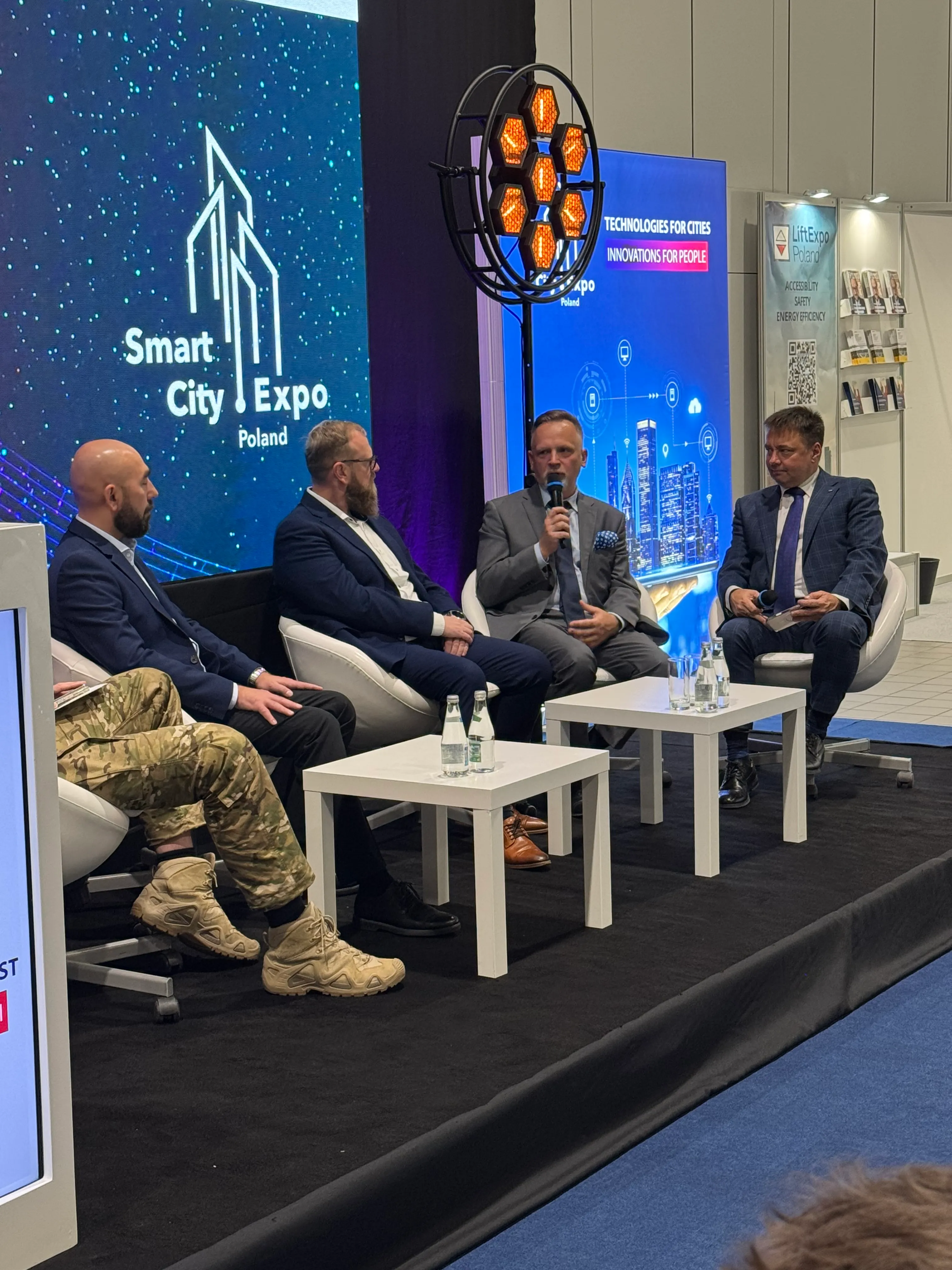 Czterech mężczyzn siedzi na scenie podczas panelu dyskusyjnego. Trzech ma garnitury, jeden mundur wojskowy i beżowe buty taktyczne. Jeden mężczyzna mówi do mikrofonu. Za nimi dwa ekrany, na jednym napis "Smart City Expo Poland", na drugim "Technologies for Cities Innovations for People". Na scenie białe stoły z butelkami wody.