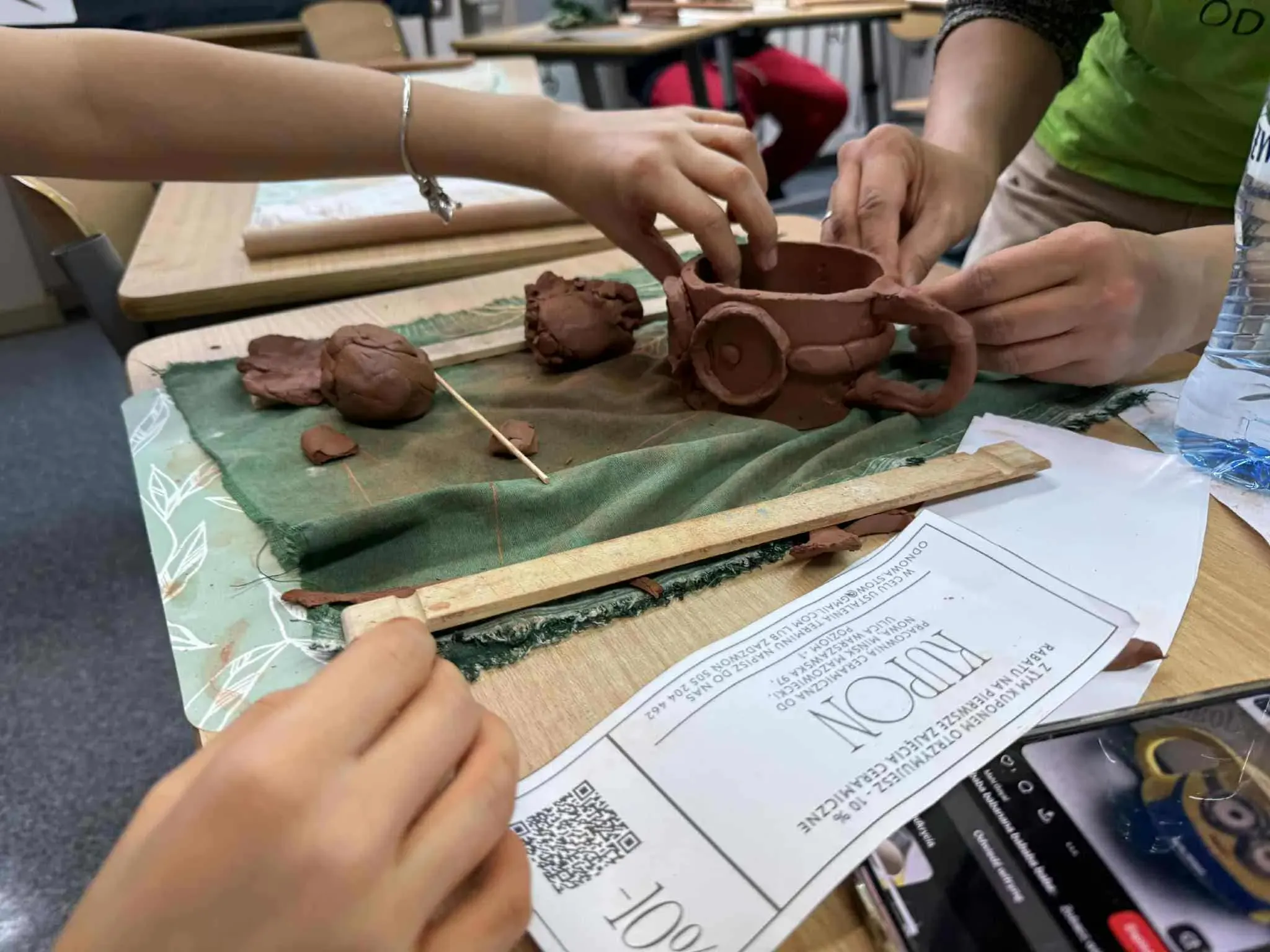 Dwie osoby modelują z gliny kubek z uchem na stole, obok leżą kawałki gliny i drewniany przyrząd. Na stole znajdują się także kartka papieru oraz butelka z wodą. Pomieszczenie jest jasne, w tle widać inne stoły i krzesła.