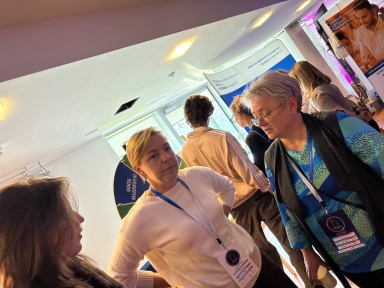 Trzy kobiety rozmawiają w jasno oświetlonym, zamkniętym pomieszczeniu podczas wydarzenia. Dwie z nich stoją przodem do siebie, trzecia jest częściowo zwrócona bokiem. Wszystkie mają przypięte identyfikatory na niebieskich smyczach. W tle stoi kilka osób i widoczne są plakaty z tekstem oraz koło z napisem „MIŃSK MAZOWIECKI”.