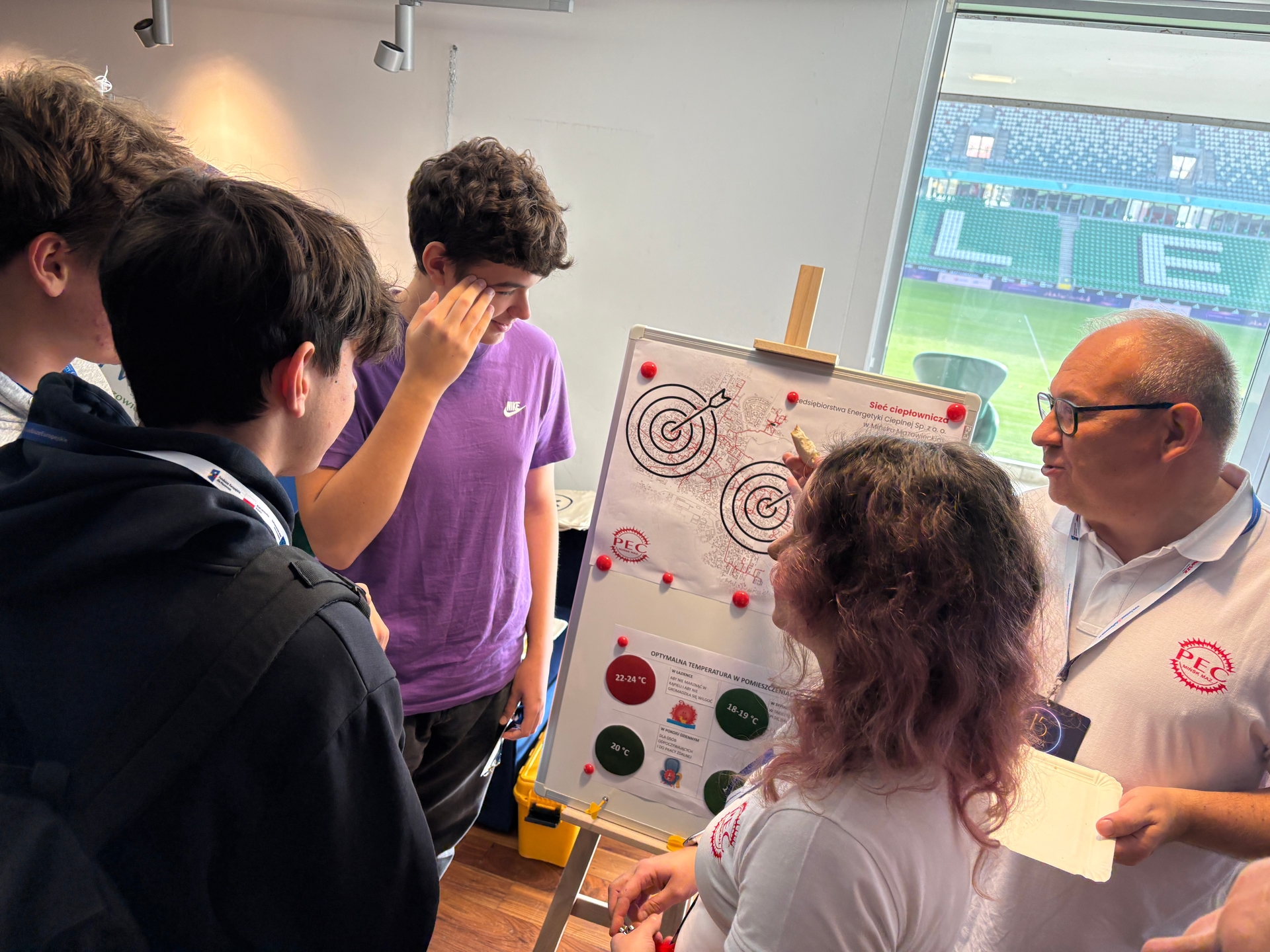 Grupa pięciu osób stoi wokół planszy z mapą i wykresami w jasnym pomieszczeniu. Dwóch młodych mężczyzn po lewej nosi ciemne ubrania, jeden w fioletowej koszulce Nike. Po prawej kobieta z kręconymi włosami i mężczyzna w białej koszulce z logo PEC wskazują na mapę. W tle widać stadion z zielonym boiskiem i trybunami.