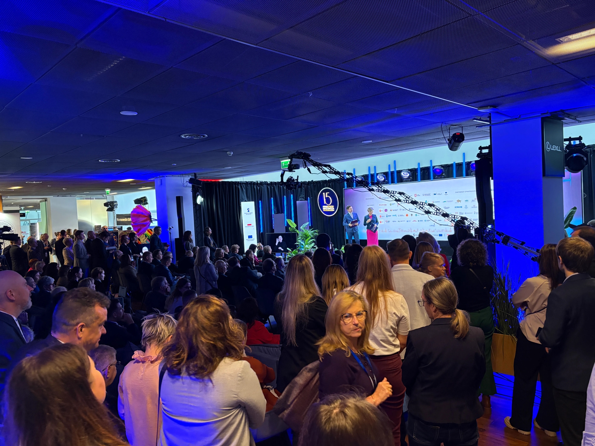 Duża grupa ludzi stoi i siedzi w hali konferencyjnej z niebieskim oświetleniem sufitowym, skupiona na dwóch prowadzących na scenie z logo "15" w tle.