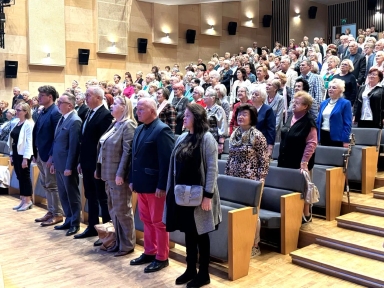 Grupa około sześćdziesięciu osób stojących w widowni jasnej, nowoczesnej sali konferencyjnej. Większość to osoby starsze, ubrani formalnie i półformalnie. Siedziska z szarymi tapicerowanymi oparciami i drewnianymi bokami. Oświetlenie ścienne na beżowych panelach.