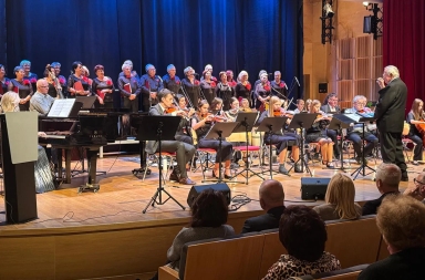 Orkiestra złożona z kilkunastu muzyków gra na różnych instrumentach strunowych i fortepianie na scenie. Za nimi stoi chór liczący około dwudziestu osób, wszyscy ubrani na czarno z czerwonymi ozdobami. Po prawej stronie stoi dyrygent prowadzący występ. Publiczność siedzi na widowni w pierwszych rzędach. Scena jest oświetlona, a tło ma ciemny, niebieski kolor.