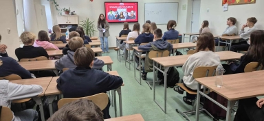 Klasa szkolna z kilkunastoma uczniami siedzącymi przy ławkach skierowanych w stronę nauczycielki na tle ściany z telewizorem i tablicą suchościeralną. Nauczycielka stoi obok ekranu z wyświetlonym napisem „Sounds and Colours of Europe”. Sala jest jasno oświetlona, na podłodze znajduje się zielony dywan.