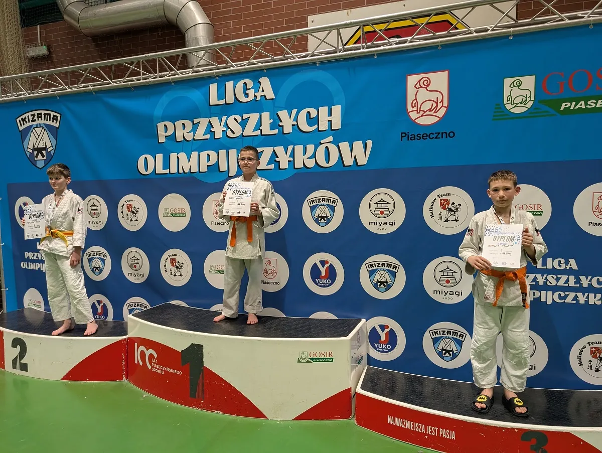 Trzech chłopców stoi na podium w hali sportowej. Chłopiec na środku stoi na najwyższym stopniu, trzyma dyplom i ma pomarańczowy pas do białego judogi. Po lewej stoi drugi zawodnik na drugim stopniu z pomarańczowym pasem i dyplomem. Po prawej stoi trzeci zawodnik na najniższym stopniu, ma judogi, pomarańczowy pas i klapki. Za nimi wisi duży niebieski baner z wieloma okrągłymi logotypami. Podłoga jest zielona.