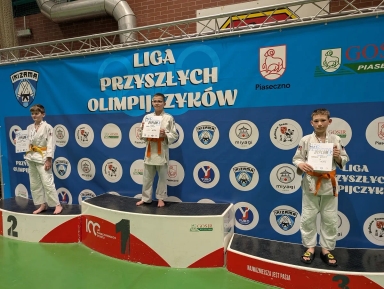 Trzech chłopców stoi na podium w hali sportowej. Chłopiec na środku stoi na najwyższym stopniu, trzyma dyplom i ma pomarańczowy pas do białego judogi. Po lewej stoi drugi zawodnik na drugim stopniu z pomarańczowym pasem i dyplomem. Po prawej stoi trzeci zawodnik na najniższym stopniu, ma judogi, pomarańczowy pas i klapki. Za nimi wisi duży niebieski baner z wieloma okrągłymi logotypami. Podłoga jest zielona.
