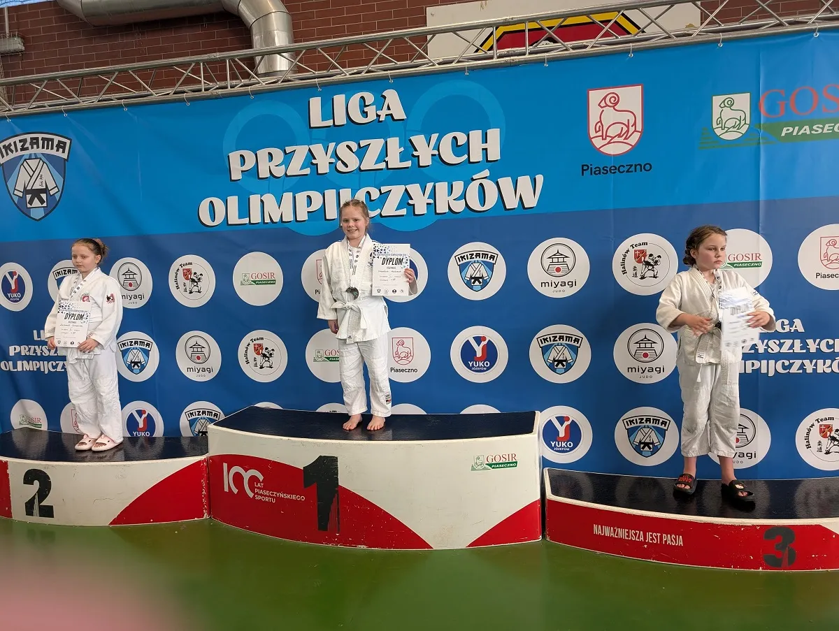 Trzy dziewczynki w białych judogach stoją na podium w hali. Centralna dziewczynka stoi na najwyższym stopniu i trzyma dyplom oraz medal. Po lewej zawodniczka stoi na drugim stopniu trzymając dokument. Po prawej zawodniczka stoi na trzecim stopniu z kilkoma kartkami w rękach. Za nimi duży niebieski baner z kilkoma okrągłymi logotypami. Podest jest czerwono-biały, podłoga zielona.