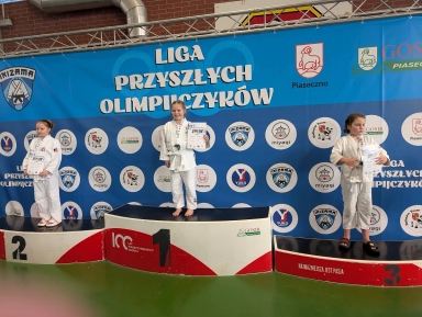 Trzy dziewczynki w białych judogach stoją na podium w hali. Centralna dziewczynka stoi na najwyższym stopniu i trzyma dyplom oraz medal. Po lewej zawodniczka stoi na drugim stopniu trzymając dokument. Po prawej zawodniczka stoi na trzecim stopniu z kilkoma kartkami w rękach. Za nimi duży niebieski baner z kilkoma okrągłymi logotypami. Podest jest czerwono-biały, podłoga zielona.