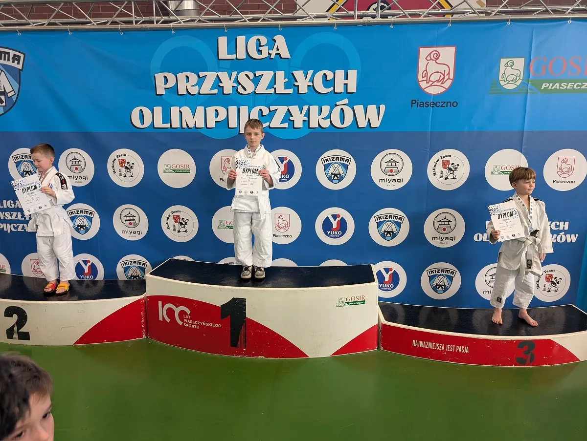 Trzech chłopców w białych judogach stoi na trzystopniowym podium w hali sportowej. Chłopiec na środku stoi na najwyższym stopniu i trzyma certyfikat. Pozostali dwaj stoją na drugim i trzecim stopniu, także trzymając certyfikaty. Za nimi jest niebieski baner z wieloma okrągłymi logotypami i jasnym tłem, a podłoga jest zielona.