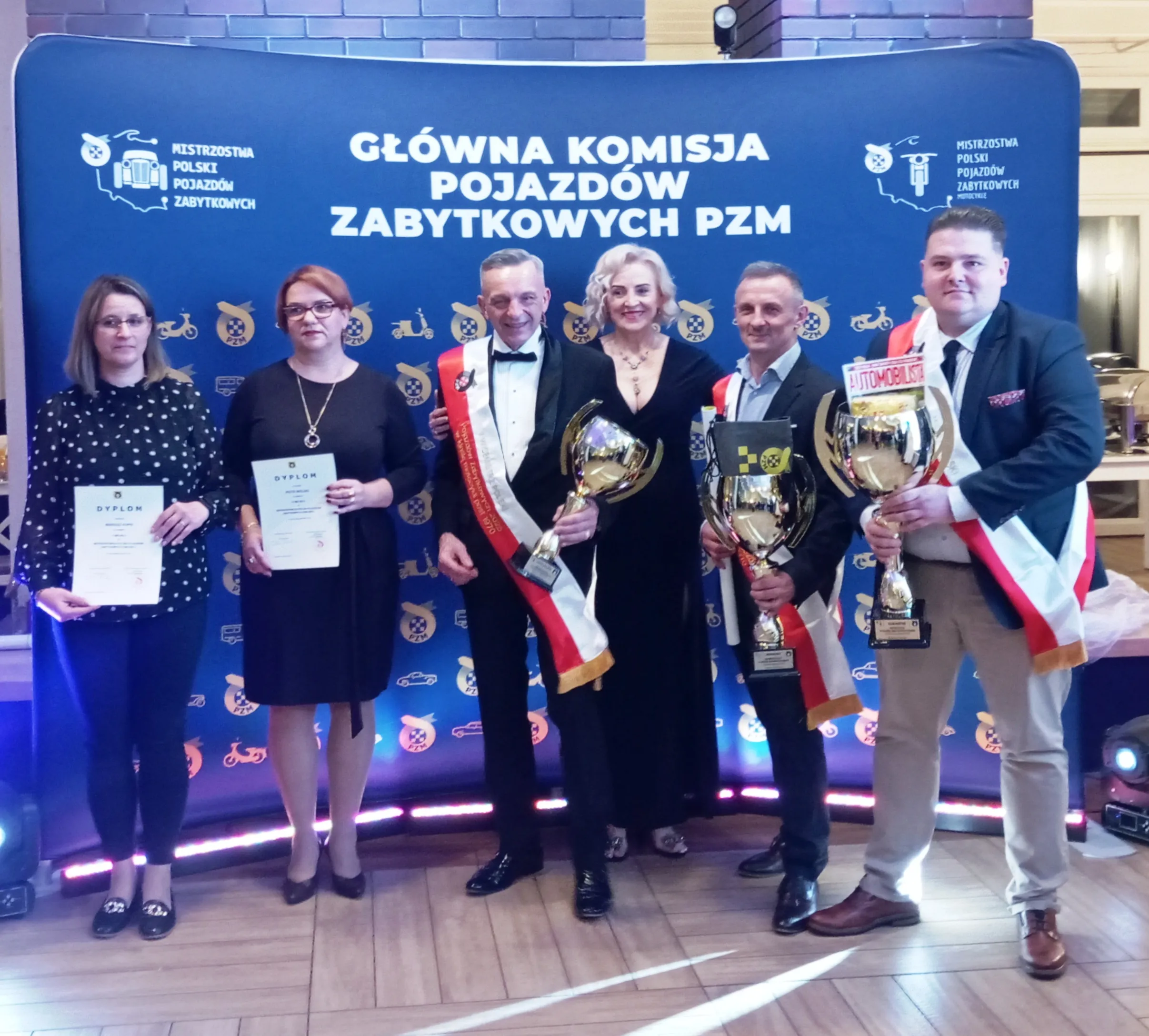 Sześć osób stojących na tle niebieskiego baneru z napisem Główna Komisja Pojazdów Zabytkowych PZM, trzy osoby z lewej trzymają dyplomy, trzy osoby z prawej trzymają puchary i mają przepaski w barwach Polski. Wnętrze z drewnianą podłogą.