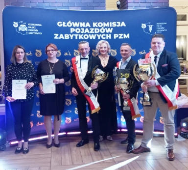 Sześć osób stojących na tle niebieskiego baneru z napisem Główna Komisja Pojazdów Zabytkowych PZM, trzy osoby z lewej trzymają dyplomy, trzy osoby z prawej trzymają puchary i mają przepaski w barwach Polski. Wnętrze z drewnianą podłogą.
