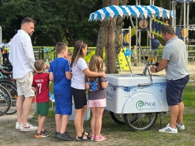 Grupa dzieci i dwóch mężczyzn stoi przy mobilnym stoisku z napojami na zewnątrz, w parku zabaw.