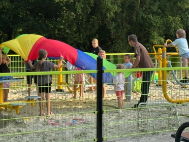 Grupa dzieci i dwóch dorosłych bawi się na piaskowym placu zabaw z ogrodzeniem, trzymając kolorowy materiał i korzystając z urządzeń na świeżym powietrzu.