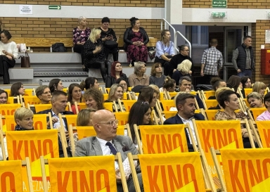 Wnętrze hali z dużą grupą dorosłych osób siedzących na żółtych, składanych krzesłach oznaczonych napisem „KINO”. W tle kilka osób siedzi na drewnianych ławkach przy ścianie z cegieł. Na ścianie widoczne zielone oznaczenie wyjścia ewakuacyjnego.