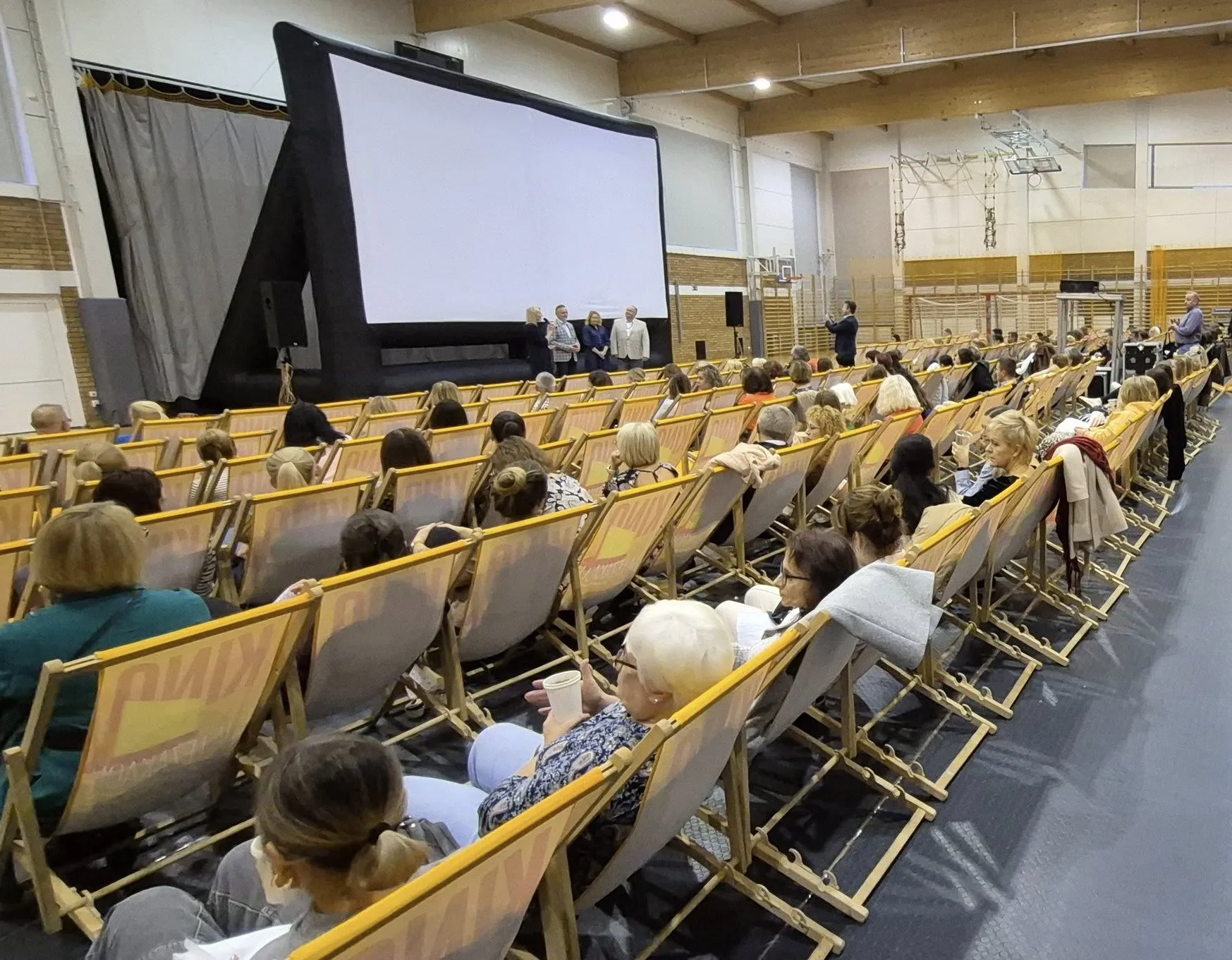 W sali gimnastycznej ustawione ustawiono rzędy leżaków z osobami siedzącymi tyłem do obserwatora. Przed nimi stoi duży biały ekran projekcyjny na czarnej ramie. Przy ekranie stoi cztery osoby. Na sali panuje sztuczne oświetlenie.