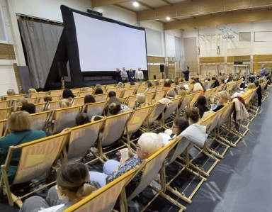 W sali gimnastycznej ustawione ustawiono rzędy leżaków z osobami siedzącymi tyłem do obserwatora. Przed nimi stoi duży biały ekran projekcyjny na czarnej ramie. Przy ekranie stoi cztery osoby. Na sali panuje sztuczne oświetlenie.