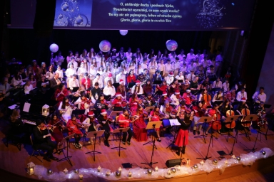 Duża grupa muzyków i chórzystów na scenie podczas koncertu świątecznego, scena udekorowana białymi i złotymi ozdobami oraz lampionami, nad zespołem wyświetlony fragment tekstu w języku polskim, muzycy grają na różnych instrumentach smyczkowych i dętych, część osób nosi czerwone czapki Mikołaja, kobieta dyryguje orkiestrą.