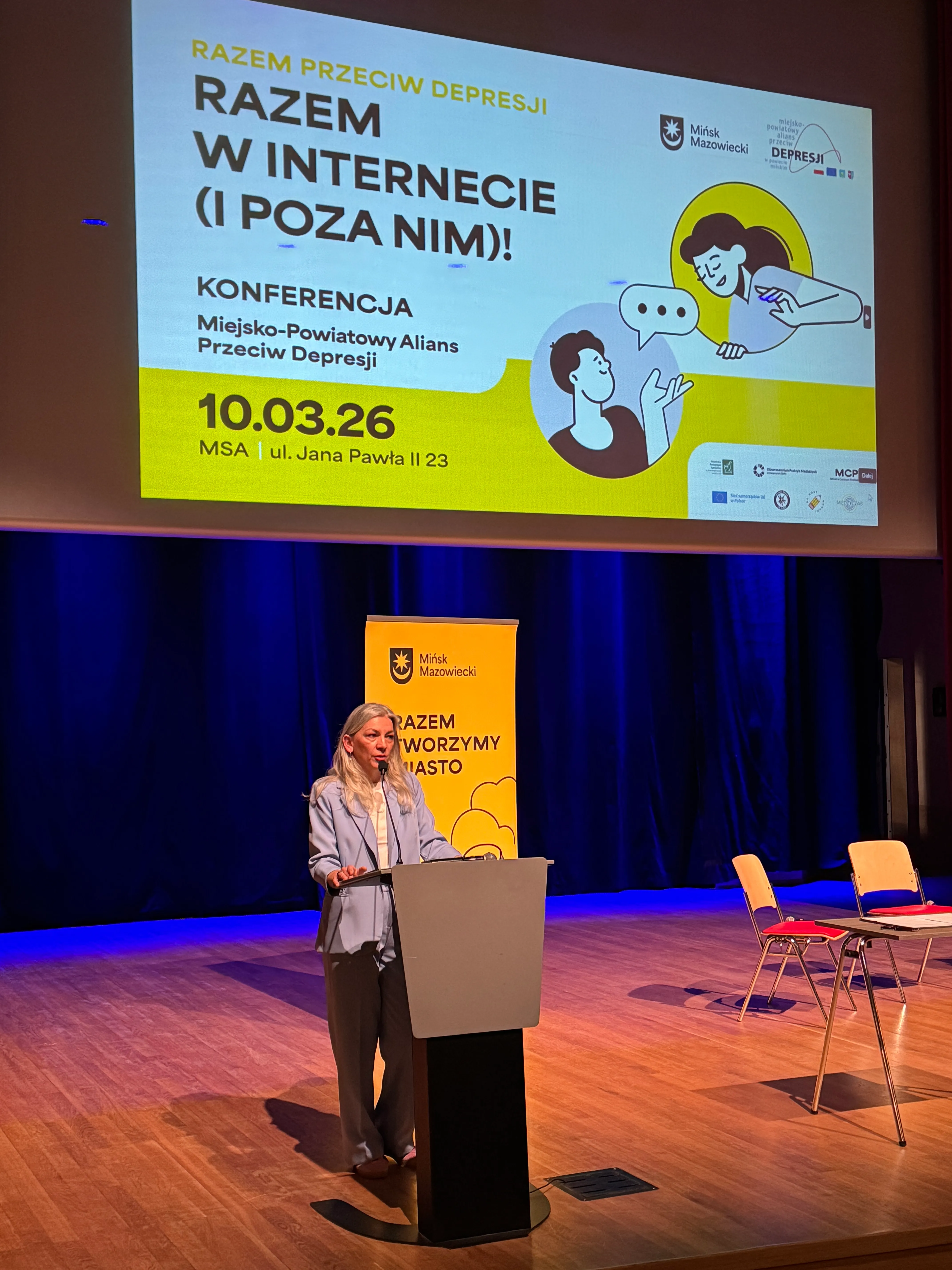 Kobieta stoi za mównicą na oświetlonej scenie w sali konferencyjnej. Nad nią wisi duży biało-żółty ekran z grafiką konferencji. Za nią stoi żółty pionowy baner z logotypem miasta. Po prawej stronie kilka pustych krzeseł przy stoliku.