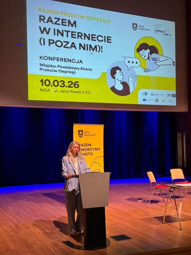 Kobieta stoi za mównicą na oświetlonej scenie w sali konferencyjnej. Nad nią wisi duży biało-żółty ekran z grafiką konferencji. Za nią stoi żółty pionowy baner z logotypem miasta. Po prawej stronie kilka pustych krzeseł przy stoliku.