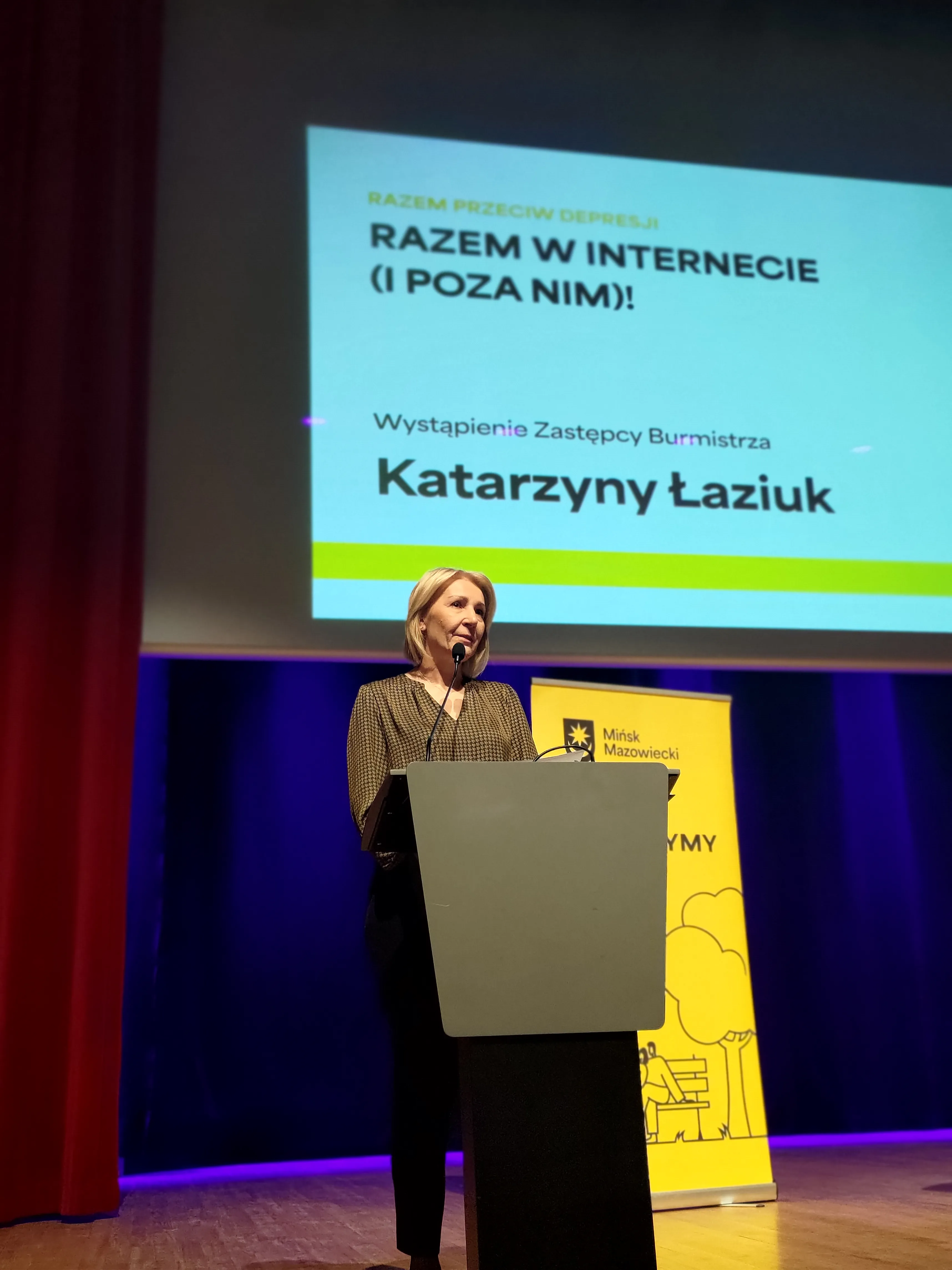 Jedna kobieta stoi za mównicą na scenie i mówi do mikrofonu. Nad nią wisi jasny ekran z projekcją. Scena ma ciemnoniebieskie tło i czerwone zasłony po lewej stronie. Po prawej stoi żółty pionowy baner z grafiką.