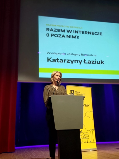 Jedna kobieta stoi za mównicą na scenie i mówi do mikrofonu. Nad nią wisi jasny ekran z projekcją. Scena ma ciemnoniebieskie tło i czerwone zasłony po lewej stronie. Po prawej stoi żółty pionowy baner z grafiką.