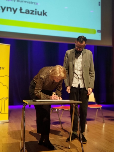 Kobieta pochyla się nad stolikiem i podpisuje dokument. Mężczyzna stoi obok niej i patrzy w dół. Scena jest na scenie z fioletowym tłem, drewnianą podłogą i krzesłami w tle. Po lewej stoi żółty pionowy baner, u góry widoczny fragment projekcji.