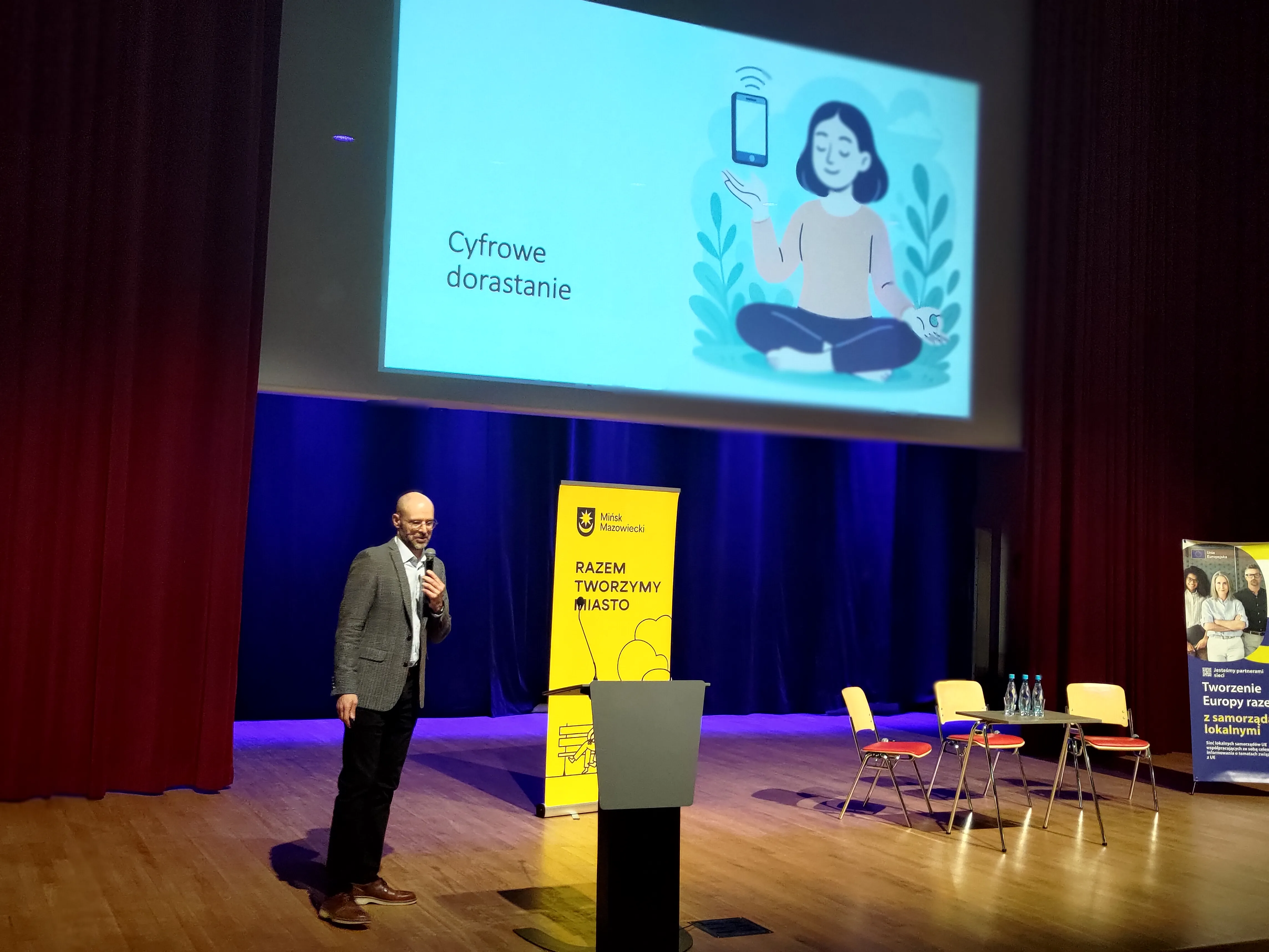 Mężczyzna stoi na scenie z mikrofonem po lewej stronie kadru. Nad sceną duży ekran z ilustracją siedzącej postaci trzymającej unoszony telefon. Za mówcą stoi pionowy żółty baner, po prawej trzy krzesła i stolik z butelkami wody. Wnętrze sali z drewnianą podłogą i ciemnoniebieską sceną oraz bordowymi kurtynami.