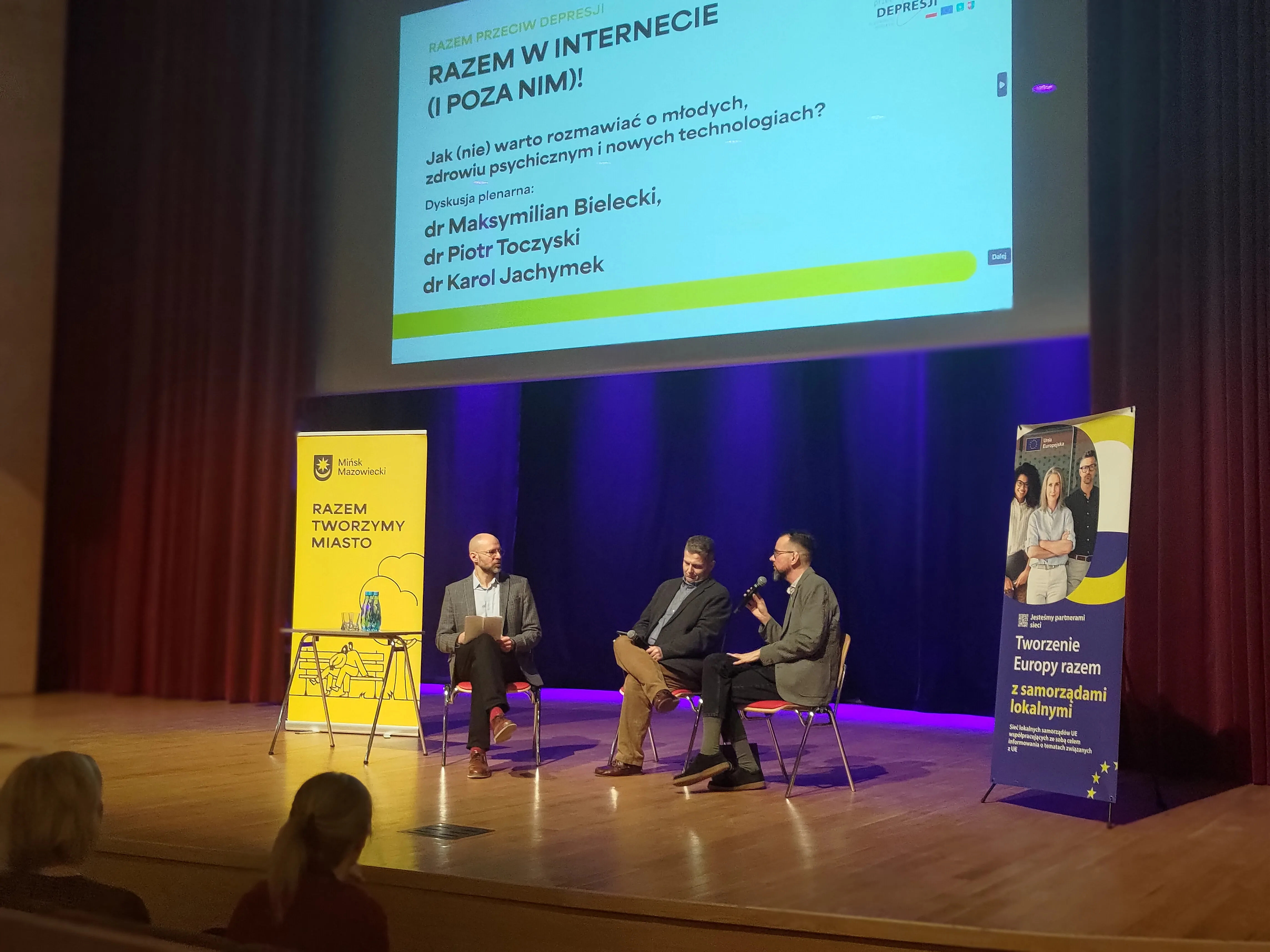 Trzech mężczyzn siedzi na scenie i rozmawia podczas panelu. Nad nimi wisi jasny ekran z prezentacją. Po obu stronach sceny stoją pionowe banery w żółto-niebieskich kolorach. Publiczność siedzi w ciemnej sali, scena oświetlona fioletowymi reflektorami.